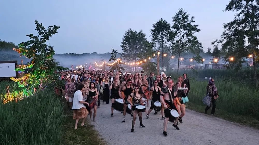 Lopende drumgroep met publiek op een festivalterrein bij avondschemer